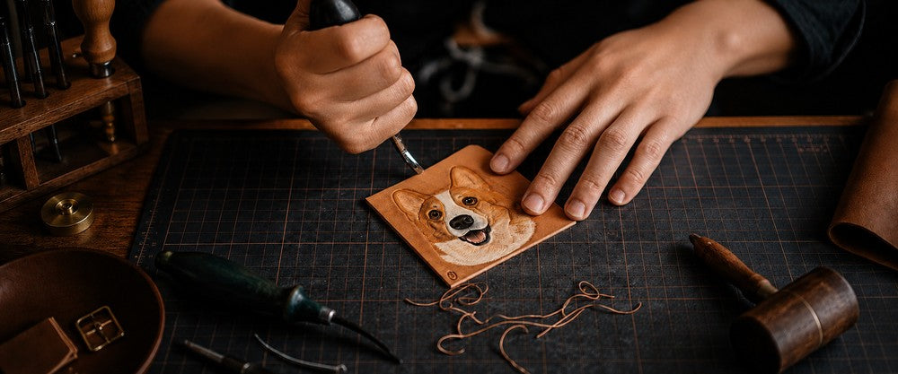 Artisan hand-carving a custom Corgi portrait into natural leather using traditional leatherworking tools on a craft workbench.