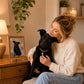Woman holding a black dog beside its personalized floral pet portrait named Luna in a cozy home.