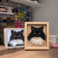 Handmade 3D felt portrait of a black and white long-haired cat displayed in a natural wood frame beside the pet’s reference photo.