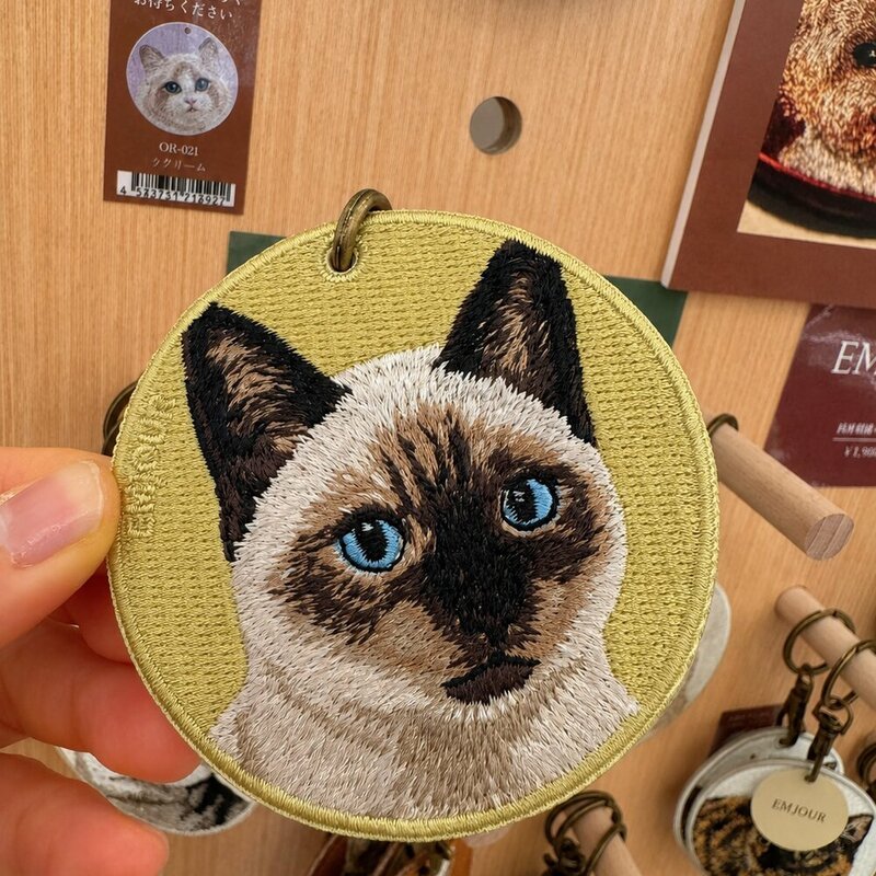Custom round embroidered cat portrait keychain featuring a blue-eyed cream-and-brown cat on an olive green fabric background.
