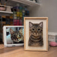 Handmade full-body 3D felt portrait of a brown tabby cat displayed in a natural wooden frame beside the cat’s reference photo.