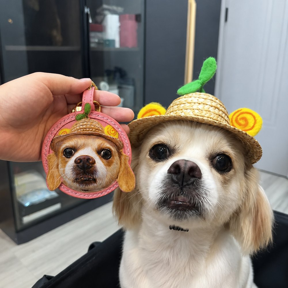 Hand holding a custom round leather keychain featuring a small dog wearing a straw hat, displayed beside the real pet in the matching hat.