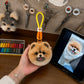 Handmade Pomeranian head bag charm with an orange hanging loop displayed beside the dog’s reference photo in an artisan workshop.