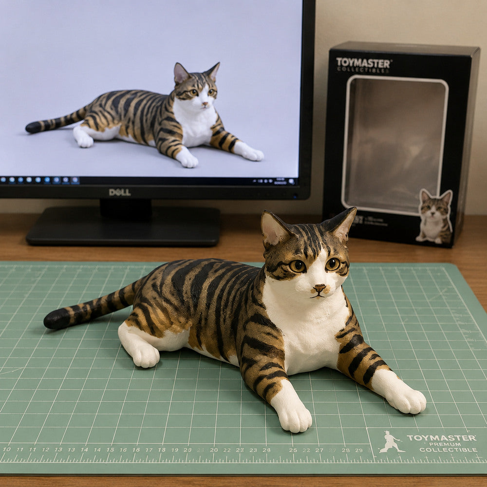 Custom brown tabby and white cat figurine in a relaxed lying pose, displayed in an artisan workshop beside its digital 3D design and collectible packaging.