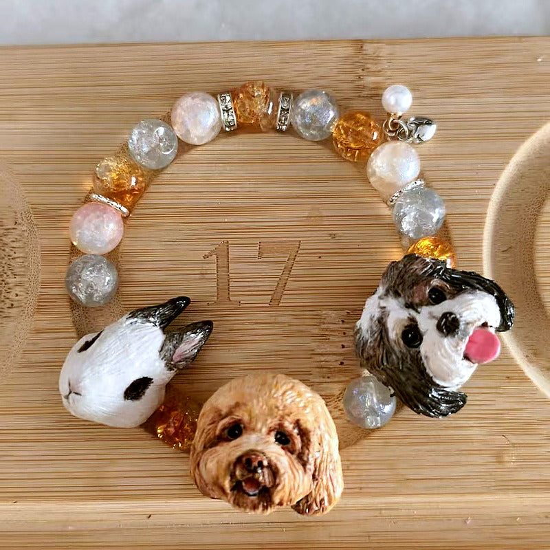 Custom three-dog portrait crystal bracelet with hand-sculpted pet face charms and clear peach beads on a bamboo board.