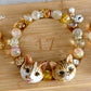Custom two-cat portrait crystal bracelet with two hand-sculpted cat face charms and warm clear beads on a bamboo board.