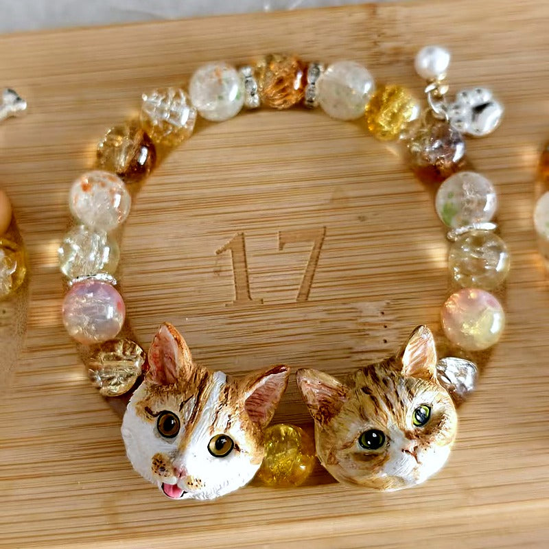 Custom two-cat portrait crystal bracelet with two hand-sculpted cat face charms and warm clear beads on a bamboo board.