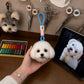 Handmade white dog head bag charm with a blue hanging loop displayed beside the pet’s reference photo in an artisan workshop.