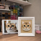 Handmade 3D felt portrait of a golden cat with large green eyes displayed in a white shadow-box frame beside its reference photo.