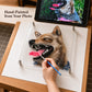 Hand-painted pet portrait in progress, with a brush adding tongue details and the dog's reference photo on a tablet.