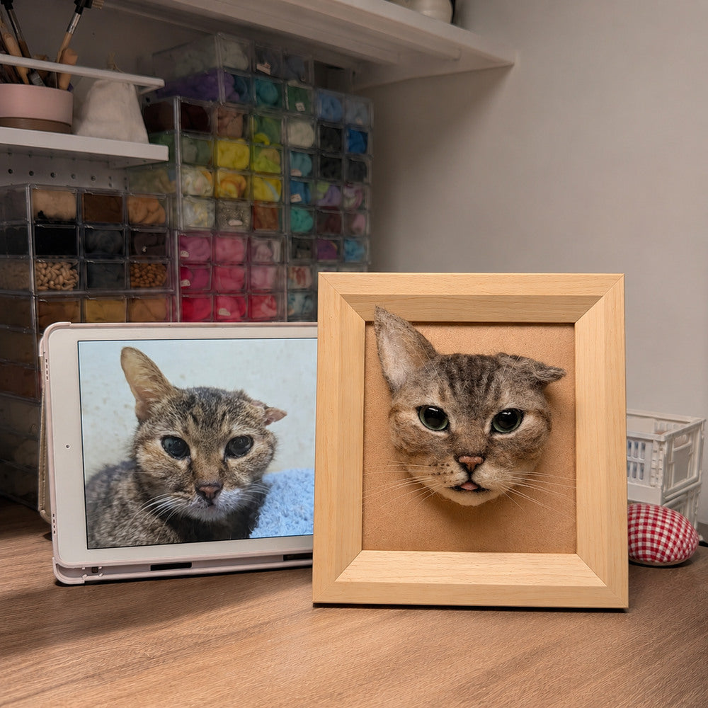 Handmade 3D felt portrait of a brown tabby cat with green eyes displayed in a natural wooden frame beside its reference photo.