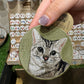 Custom round embroidered gray tabby cat portrait keychain with an olive green fabric background and antique brass-tone ring.