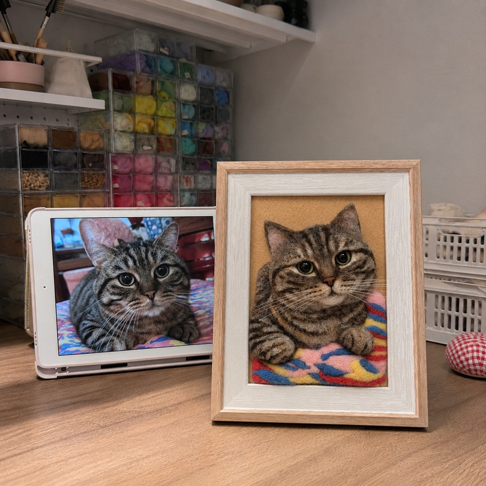 Handmade 3D felt portrait of a brown tabby cat resting on a colorful blanket, displayed in a wooden frame beside its reference photo.