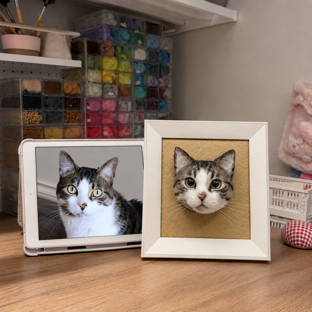Handmade 3D felt portrait of a gray and white tabby cat displayed in a white shadow-box frame beside the cat’s reference photo.