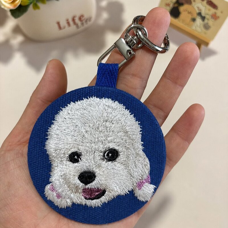 Custom round embroidered white dog portrait keychain on a royal blue fabric background with a silver-tone swivel clasp.